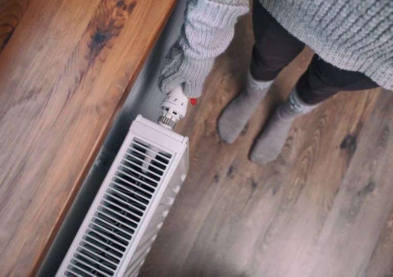Person wearing gray sweater, gloves, and socks adjusts the temperature knob on a white radiator on a wooden floor.