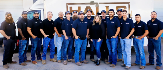 A group of sixteen people in matching black shirts and jeans pose in front of a truck with the word "HALLS" on it.