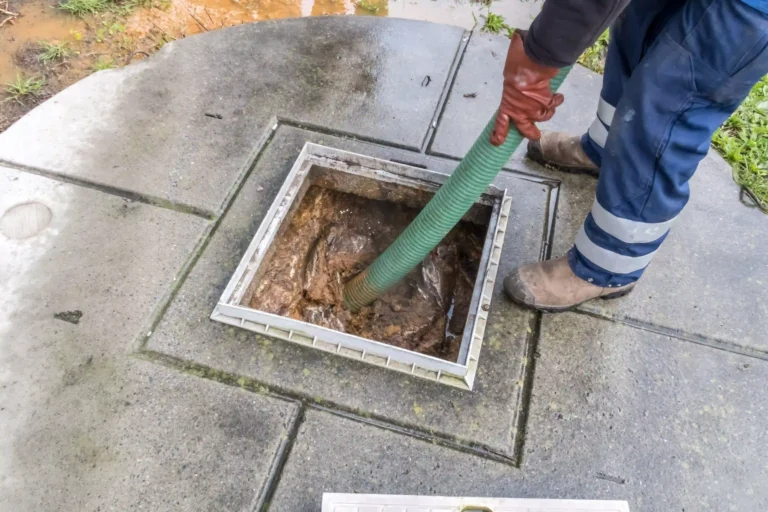 A Halls Rooter & Plumbing team member in gloves, boots, and reflective pants uses a green hose to clean a dirty drain on a concrete surface—an important step before performing a Sewer Camera Inspection in Midland & Odessa, TX.
